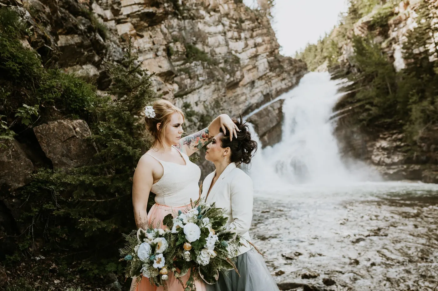 Two brides forehead to forehead before a mountain waterfall