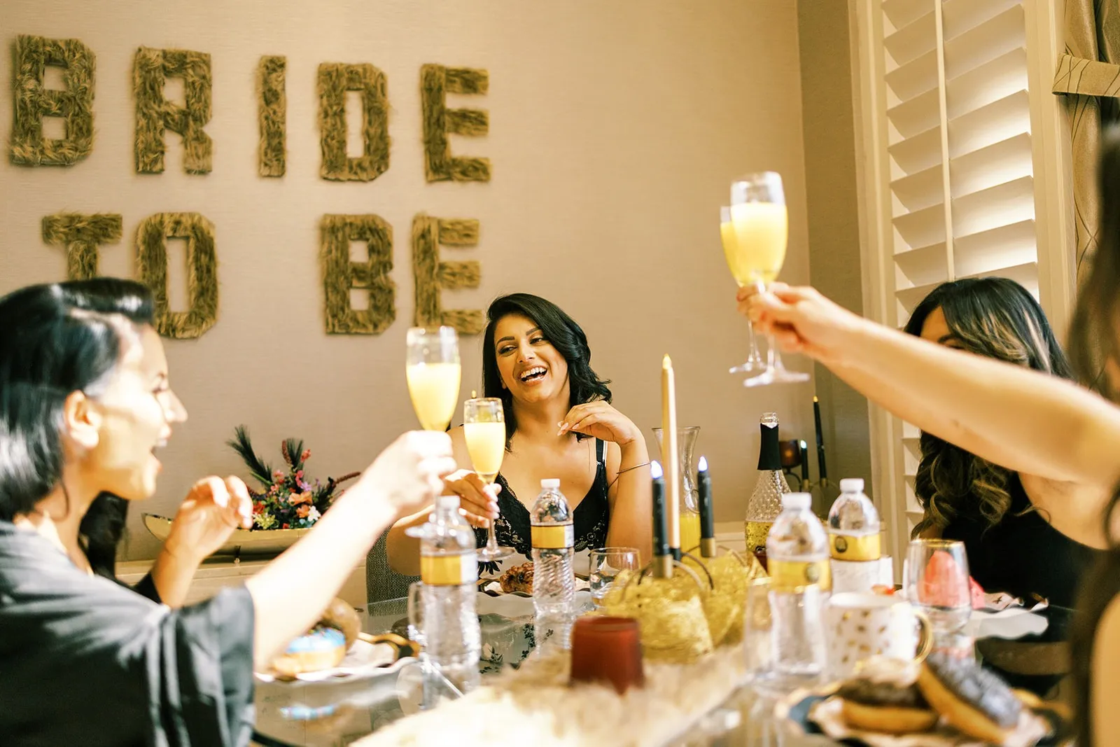 Friends raising mimosas at a bridal brunch beneath a moss 'Bride to Be' sign